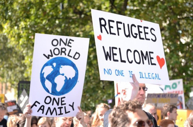 Public demonstrators hold up signs that say "one world, one family" and "refugees welcome; no one is illegal"