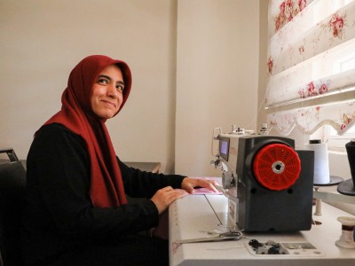 An image of a woman and small business owner sitting in front of a sewing machine and smiling at the camera
