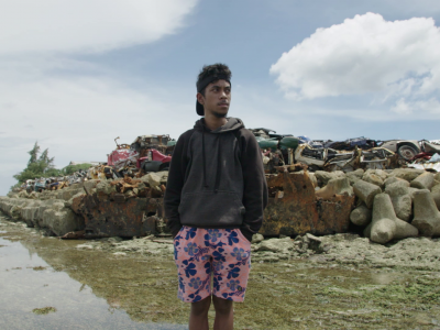 Teenage boy stands infront of a constructed sea wall made of old cars and blocks of cement